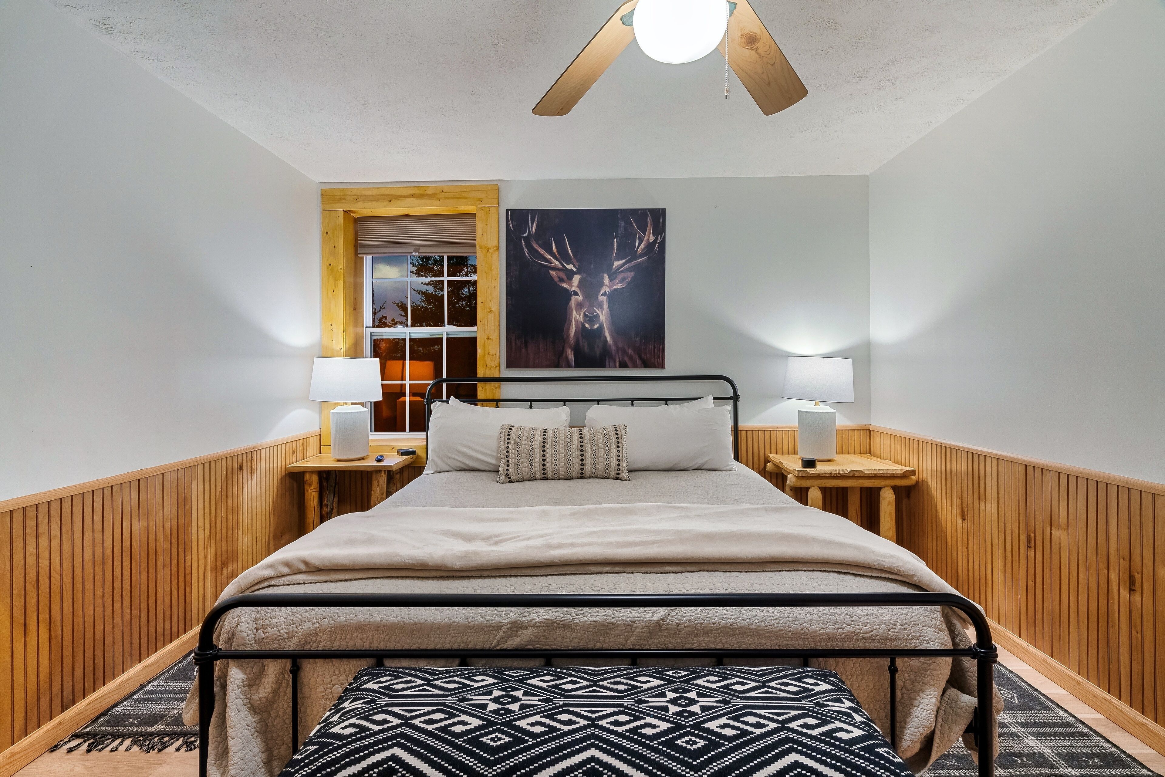 A bedroom with a bed, bedside lamps, a wooden headboard, a window with blinds, and a painting of a deer on the wall.