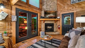 A cozy cabin interior with a stone fireplace, wooden walls, and a large window overlooking a forest. - Stunning Sunset Views! Hot Tub, 2 Fire Pits, Game Loft, Dogs Welcome, Pool Open! (Sevierville)