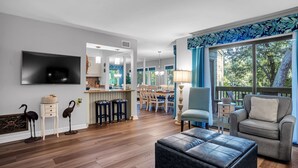 A spacious living room with a flat-screen TV, a dining area with wooden chairs, and a large window with blue curtains. - 2316 Racquet Club at The Sea Pines Resort (Hilton Head Island)