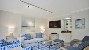 Un salon comprenant un canapé, une table basse en verre, des fauteuils en osier, un meuble blanc et un téléviseur à écran plat.