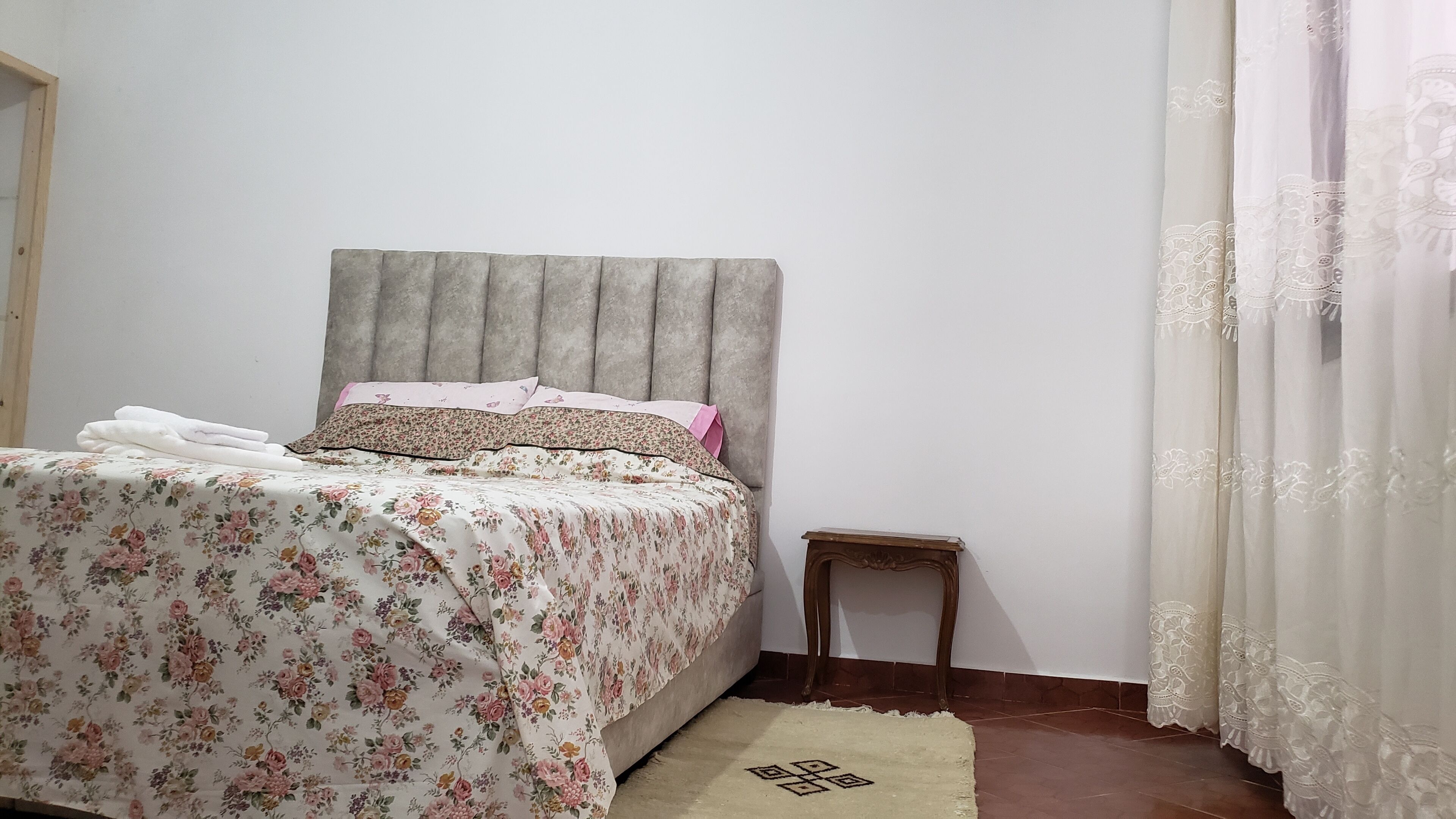A bedroom with a bed, a small wooden nightstand, a patterned bedspread, and a plain wall.