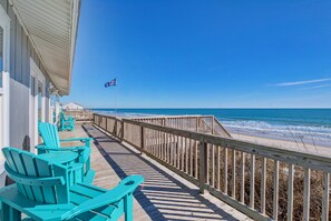 A wooden deck with turquoise Adirondack chairs, a wooden railing, and a view of the ocean.