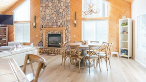 A cozy cabin interior with a stone fireplace, wooden walls, and a dining area with multiple chairs. - Spacious cabin with game room free WIFI & parking (Shaver Lake)
