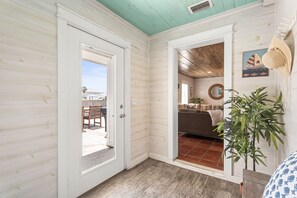 A room with a wooden floor, a door leading to an outdoor area, a plant, and a wall with a circular mirror and a framed picture.