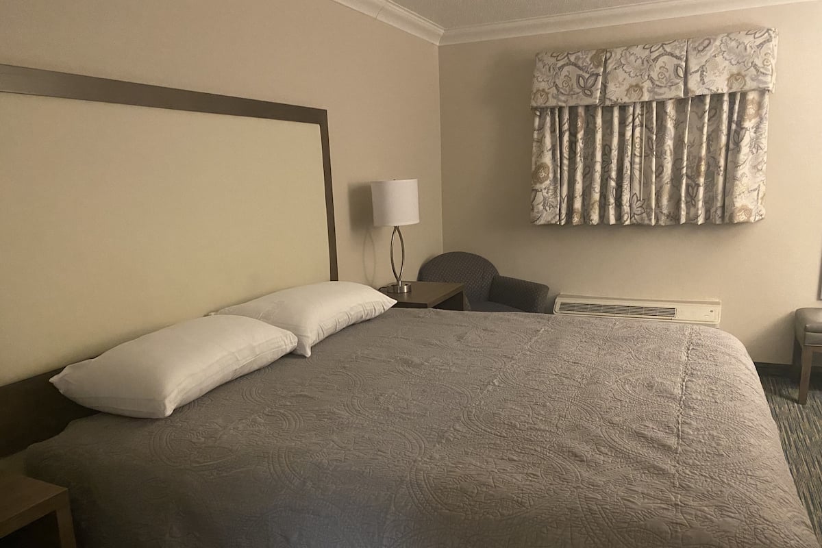 Ein Hotelzimmer mit einem großen Bett, einem Nachttisch mit Lampe, einem Sessel und einem Vorhangfenster.