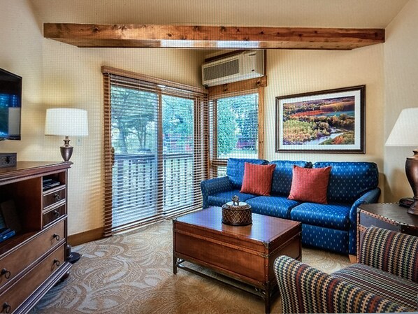 A living room with a blue sofa, a wooden coffee table, a patterned armchair, a TV on a wooden cabinet, and a painting on the wall. - Perfect Studio Condo Resort (Galena)