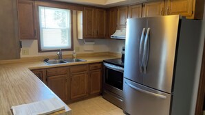 Una cocina con armarios de madera, un refrigerador de acero inoxidable, un lavamanos doble y una ventana con persianas.