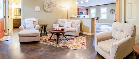 A spacious living room with a large sofa, a coffee table, and a patterned rug. There is a kitchen area in the background with a bar and a dining table.