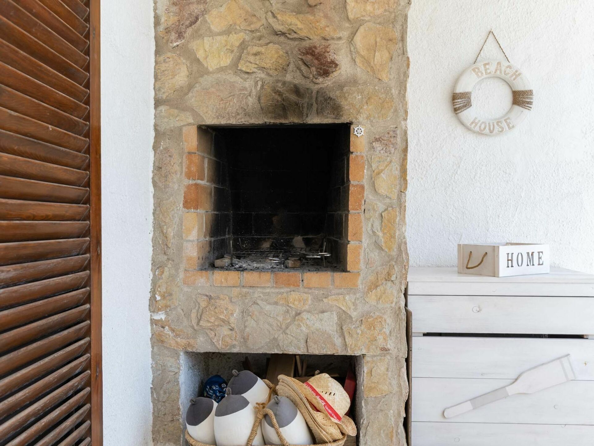 A stone fireplace with a log storage area below, a white wall with a 'BEACH HOUSE' sign, and a wooden door with horizontal slats.