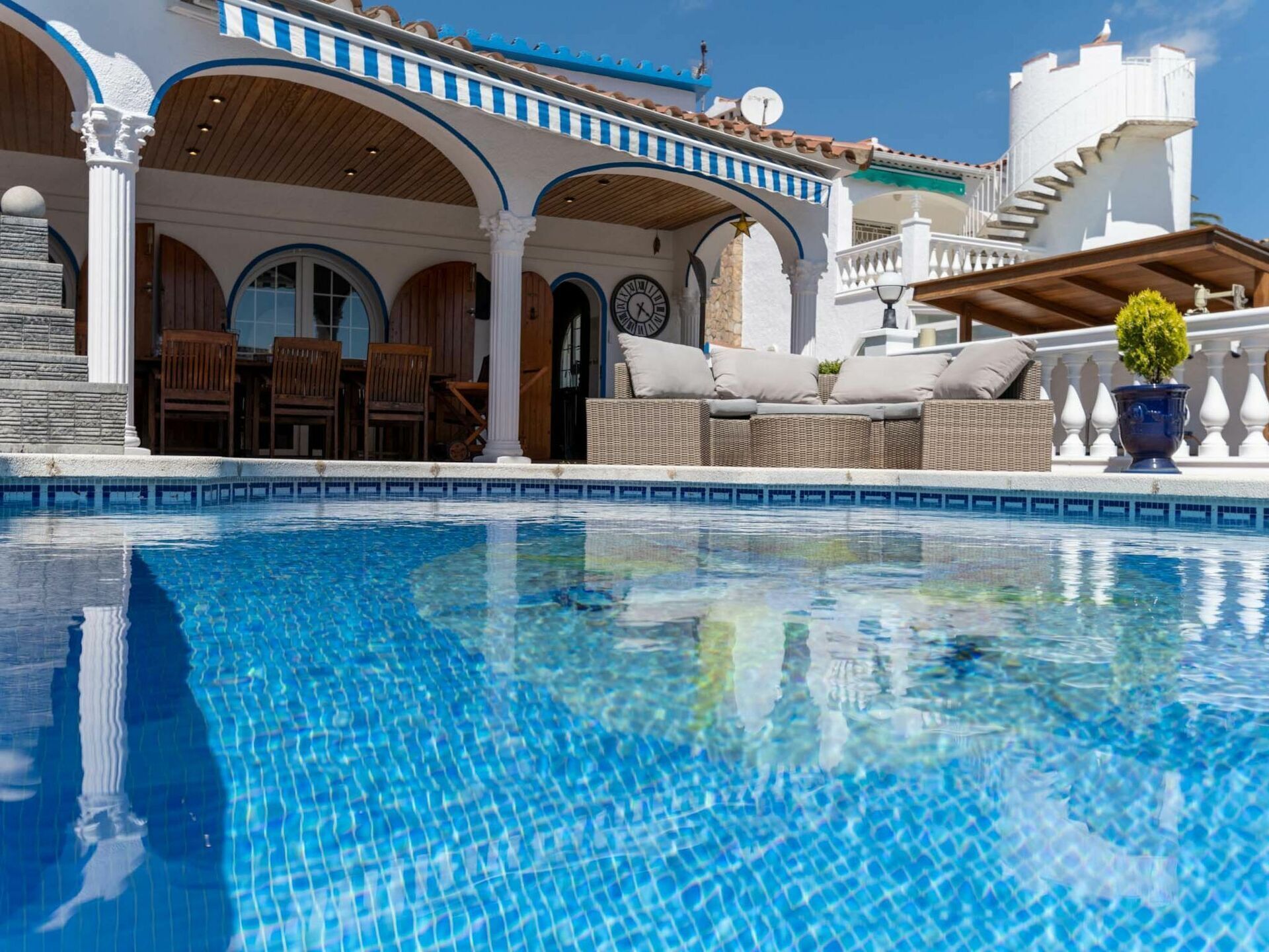 A pool area with a tiled edge, outdoor seating, and a building with arches and a clock.