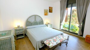 Une chambre à coucher comprenant un lit, un banc, une table de chevet, une fenêtre avec des rideaux et un mur orné de deux tableaux.