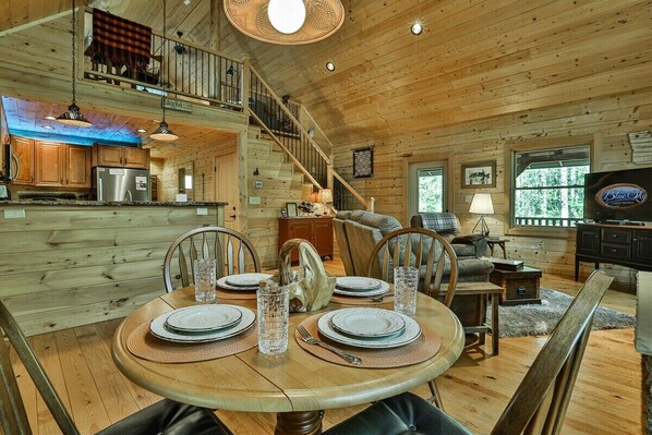 Interior de una cabaña de madera con una mesa de comedor para cuatro, un área de cocina, una sala de estar con sofá y una escalera.