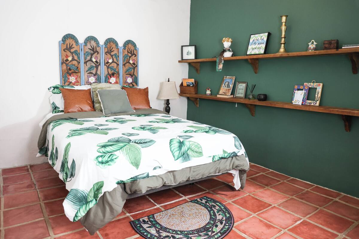 Un dormitorio con cama, almohadas, una mesita de noche, lámpara, un adorno en la pared y estantes de madera con diversos objetos.