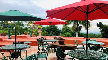 An outdoor patio area with round tables and chairs, red and green umbrellas, and a view of the landscape.