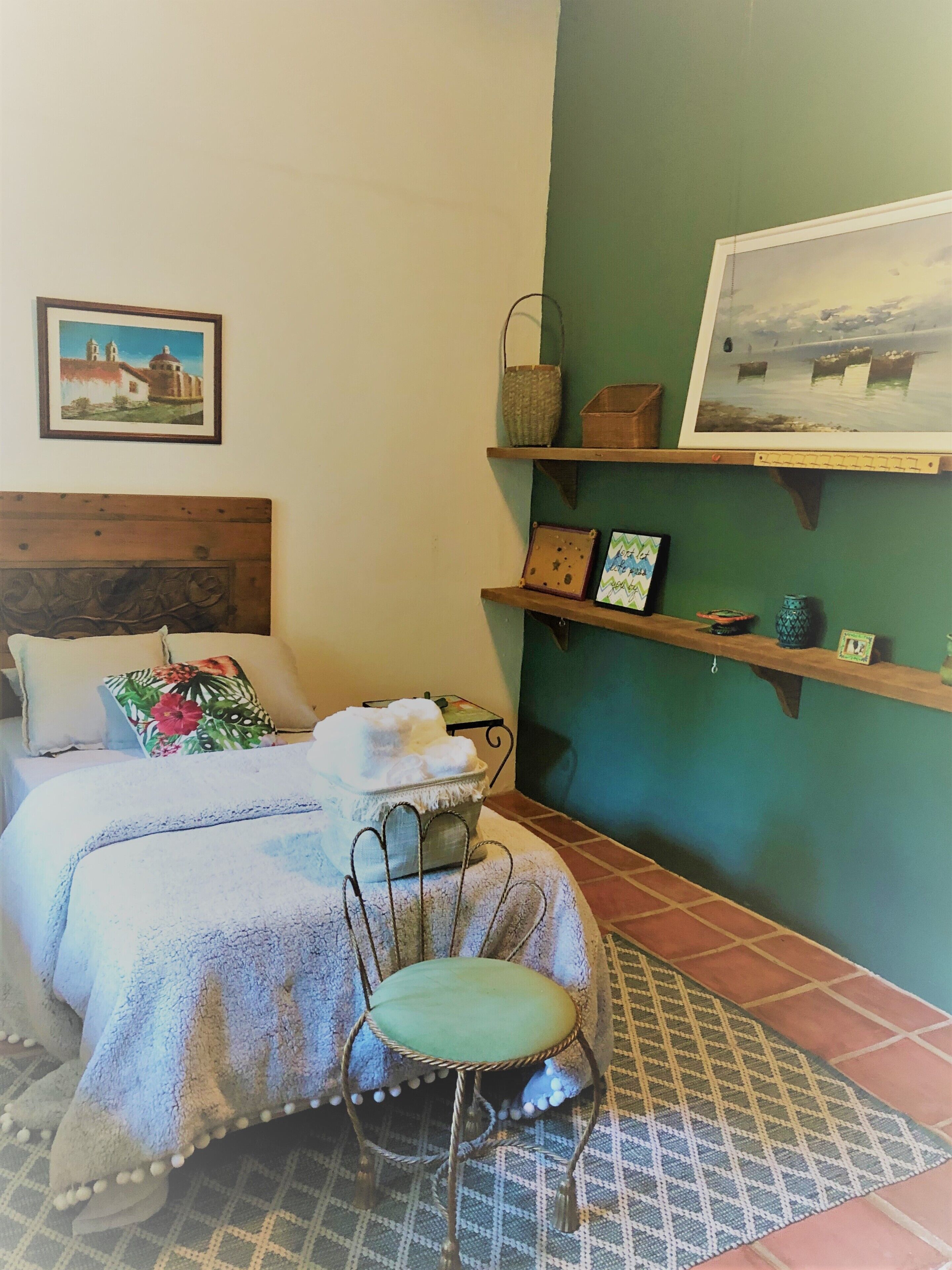 A bedroom with a bed, a chair, a nightstand, a shelf with decorative items, and a painting on the wall.