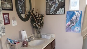 Une salle de bain avec un meuble-lavabo en bois, des toilettes, un lavabo, un miroir et divers éléments de décoration murale, notamment des affiches et une horloge.