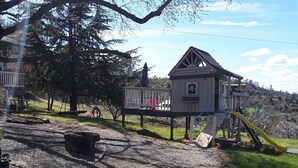 Une aire de jeux avec un toboggan et des balançoires, une petite structure en bois et un arbre en arrière-plan.