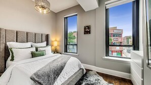 A bedroom with a large bed, a headboard, a nightstand, a lamp, and a view of the city through large windows. - Lovely 4 Bedroom Spot located in Charming Old Town (Chicago)