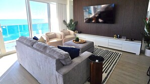 Ein modernes Wohnzimmer mit grauem Sofa, einem Flachbildfernseher an einer Holzwand und einem Balkon mit Blick auf den Ozean.