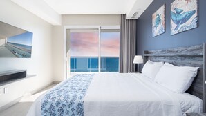 Una habitación de hotel con una cama grande, un televisor sobre un mueble, una mesita de noche y vista al océano a través de un ventanal.