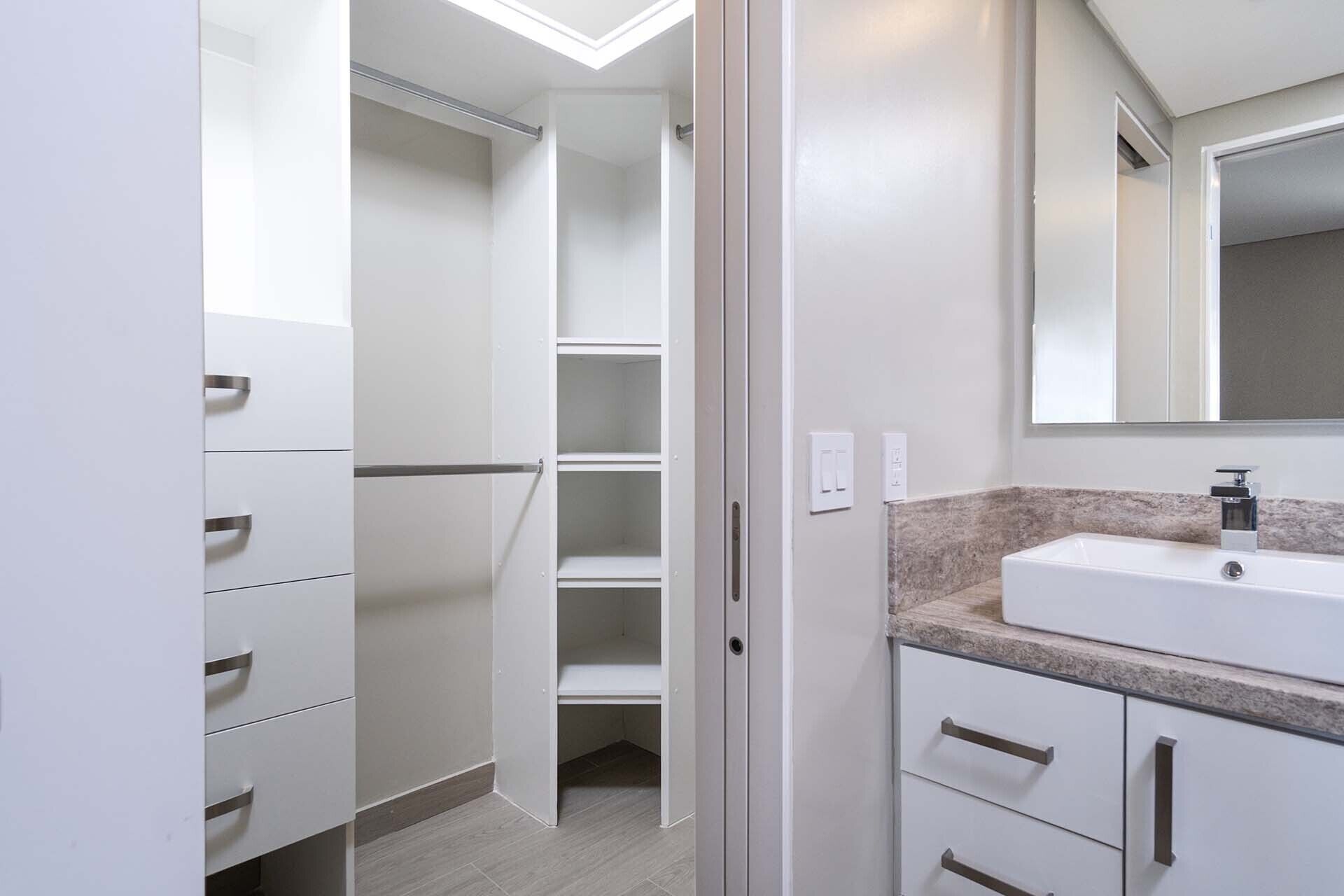 A modern bathroom with a built-in sink, a mirror, and a walk-in closet with shelves and drawers.