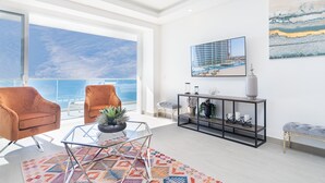 A modern living room with a large window overlooking a body of water, a glass coffee table, a sofa, and a console table with decorative items. - Amazing 2 bedroom Beach Front Condominium at Encantame C1102 (Puerto Peñasco)