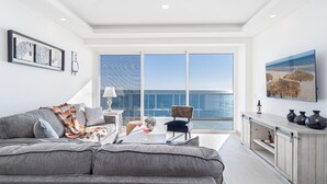 Een moderne woonkamer met een grijze bank, een bijzettafeltje met een lamp, een stoel met een kussen met luipaardprint, en een groot raam met uitzicht op de zee.