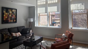 Un salon avec un canapé noir, une table basse et un fauteuil. De grandes fenêtres sont équipées de stores, et on y trouve une lampe ainsi qu’une œuvre d’art encadrée accrochée au mur.
