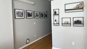 Un couloir avec, sur le mur de gauche, une série de photographies en noir et blanc encadrées, une grille d’aération blanche, et, sur le mur de droite, une rangée d’étagères à cadre noir où sont exposées des plantes en pot et des objets décoratifs.