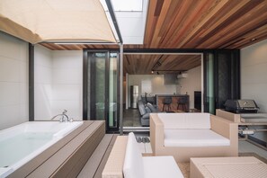 A modern bathroom with a large bathtub, wooden flooring, and a glass door leading to a living area with a sofa and a grill. - Villa Kanasa YUKUI (Onna)