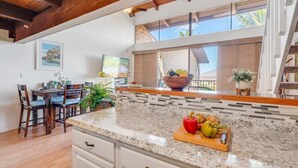 A kitchen with a granite countertop, wooden chairs, a dining table, and a window with a view of palm trees.