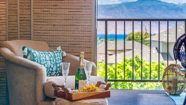 A balcony with a view of mountains and greenery, a sofa with a bottle and glasses, and a wicker basket with lemons.