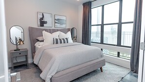 Une chambre à coucher moderne avec un grand lit, deux tables de chevet, une commode, une fenêtre avec des rideaux et des œuvres d’art accrochées au mur.