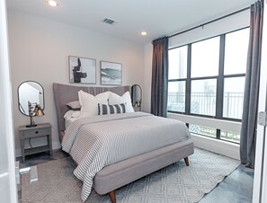 A modern bedroom with a large bed, two bedside tables, a nightstand, a window with curtains, and artwork on the wall.