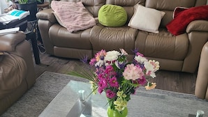 Un soggiorno con un divano in pelle marrone, un tavolino da caffè in vetro con un vaso di fiori e una piccola pianta verde su un tavolino laterale.