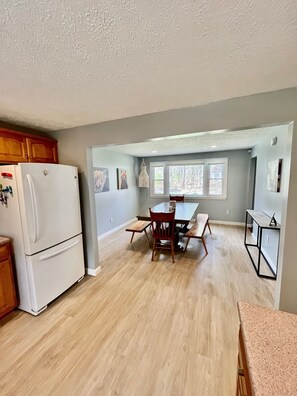 A kitchen with a white refrigerator, wooden cabinets, a dining table with chairs, and a window with a view of trees.