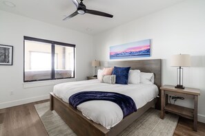 Un dormitorio con cama, ventilador de techo, ventana, mesita de noche, lámpara y un cuadro en la pared.
