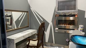 A bathroom with a marble vanity, a wooden chair, a mirror with a gold frame, a built-in fireplace, and a blue sink.