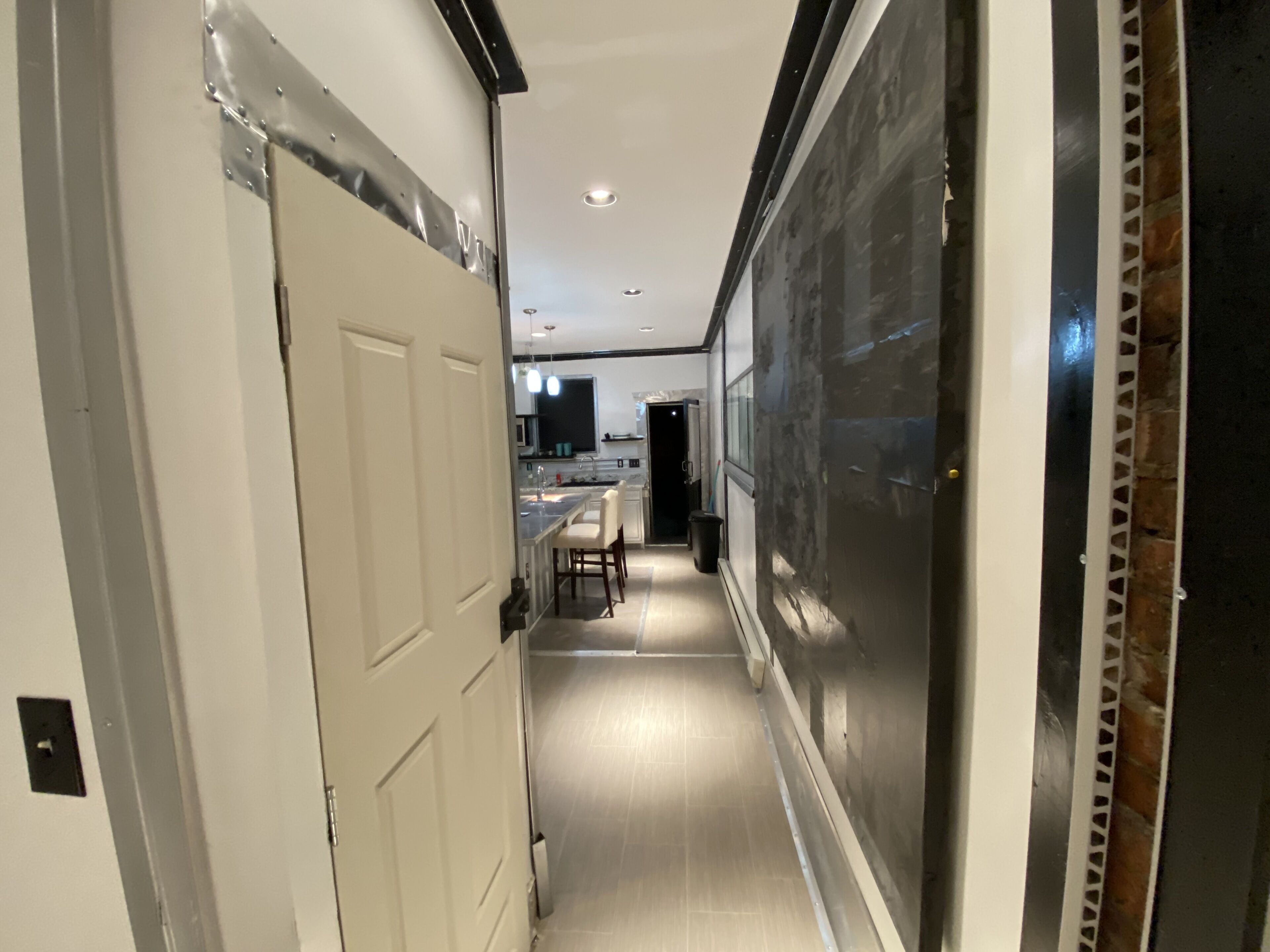 Un pasillo estrecho con una puerta blanca a la izquierda, una zona de cocina con mostrador y sillas en el centro, y un ventanal grande a la derecha.