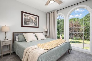 A bedroom with a large window, a bed with a quilt, a nightstand with a lamp, and a framed picture on the wall.