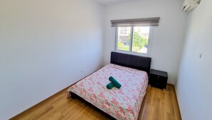 A bedroom with a bed, wooden flooring, a window with blinds, and an air conditioning unit. - Springfield Park Apartment (Larnaca)