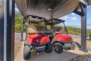Deux voiturettes de golf rouges sont garées sous un abri, avec un chemin de gravier et de la verdure en arrière-plan.