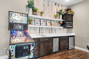 A kitchen with a pinball machine, dark wooden cabinets, stainless steel appliances, and a white countertop.