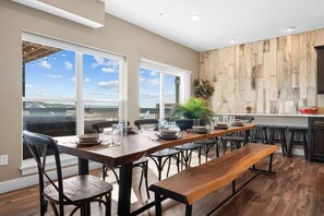 Uma área de refeições com uma mesa de madeira comprida, várias cadeiras e um banco. Grandes janelas oferecem vista para o exterior.