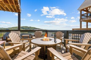 An outdoor seating area with a fire pit, wooden chairs, and a view of a lake and hills.