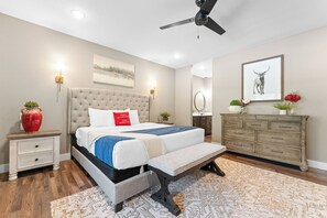 A bedroom with a bed, bedside tables, a bench, a dresser, a ceiling fan, and a painting on the wall.