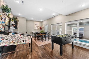 Sala de estar moderna com mesa de futebol, área de jantar com mesa redonda e cadeiras, e uma grande janela com vista.
