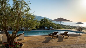 A pool area with lounge chairs and umbrellas, surrounded by trees and a view of the sea and mountains.