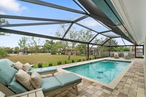 A covered outdoor swimming pool area with lounge chairs and a view of a grassy yard.