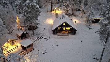 A snow-covered cabin with a lit interior, surrounded by trees and a snow-covered ground.